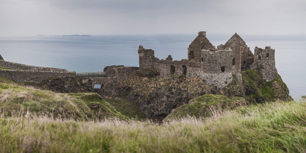 Grey blue Skies with ancient stone building sitting on the edge. Green, tall grass in the foreground 