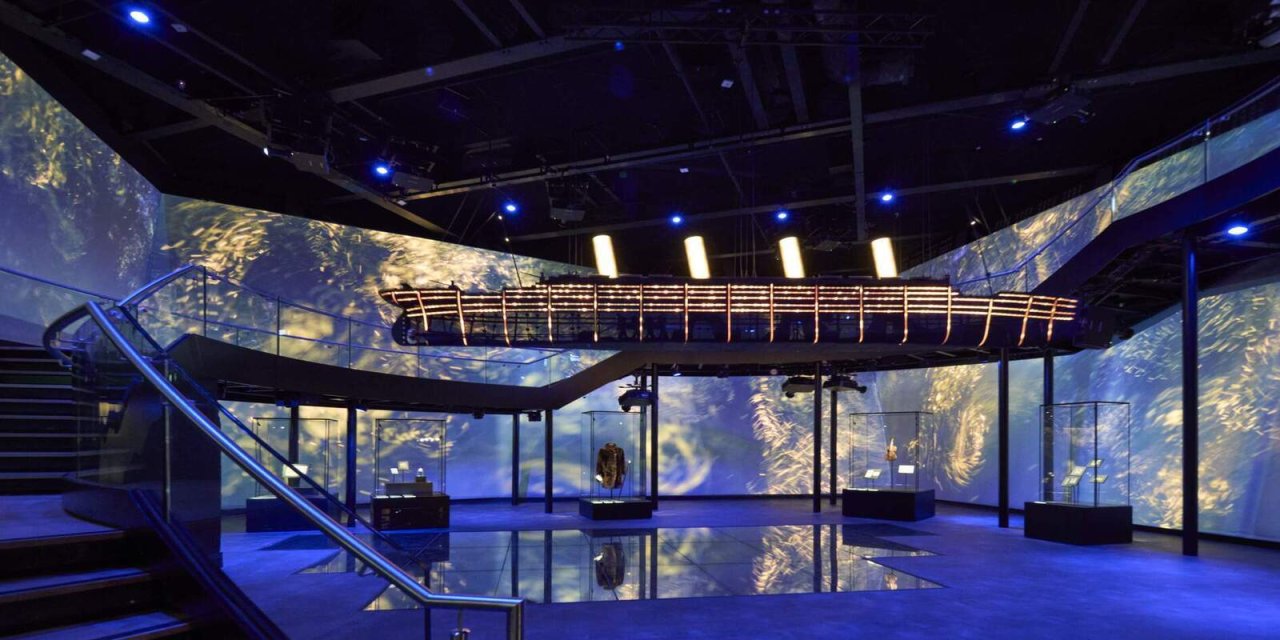 Blue light illusions projected on the 3 back walls. stairs with silver hand rail from the left foreground all the way around the room. 5 clear glass boxes with artefacts on the ground in the middle of the room. 