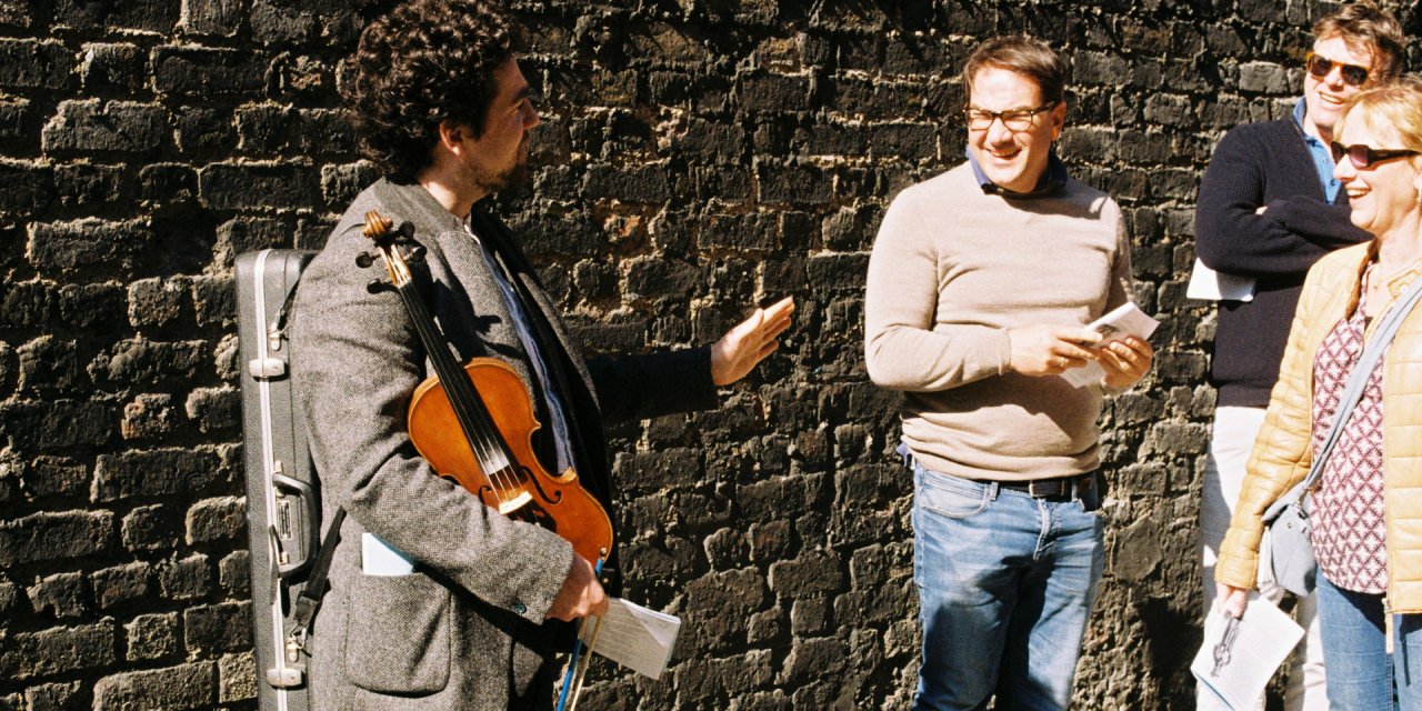 Man wearing grey suit holding violin standing outside at brick wall giving a music tour